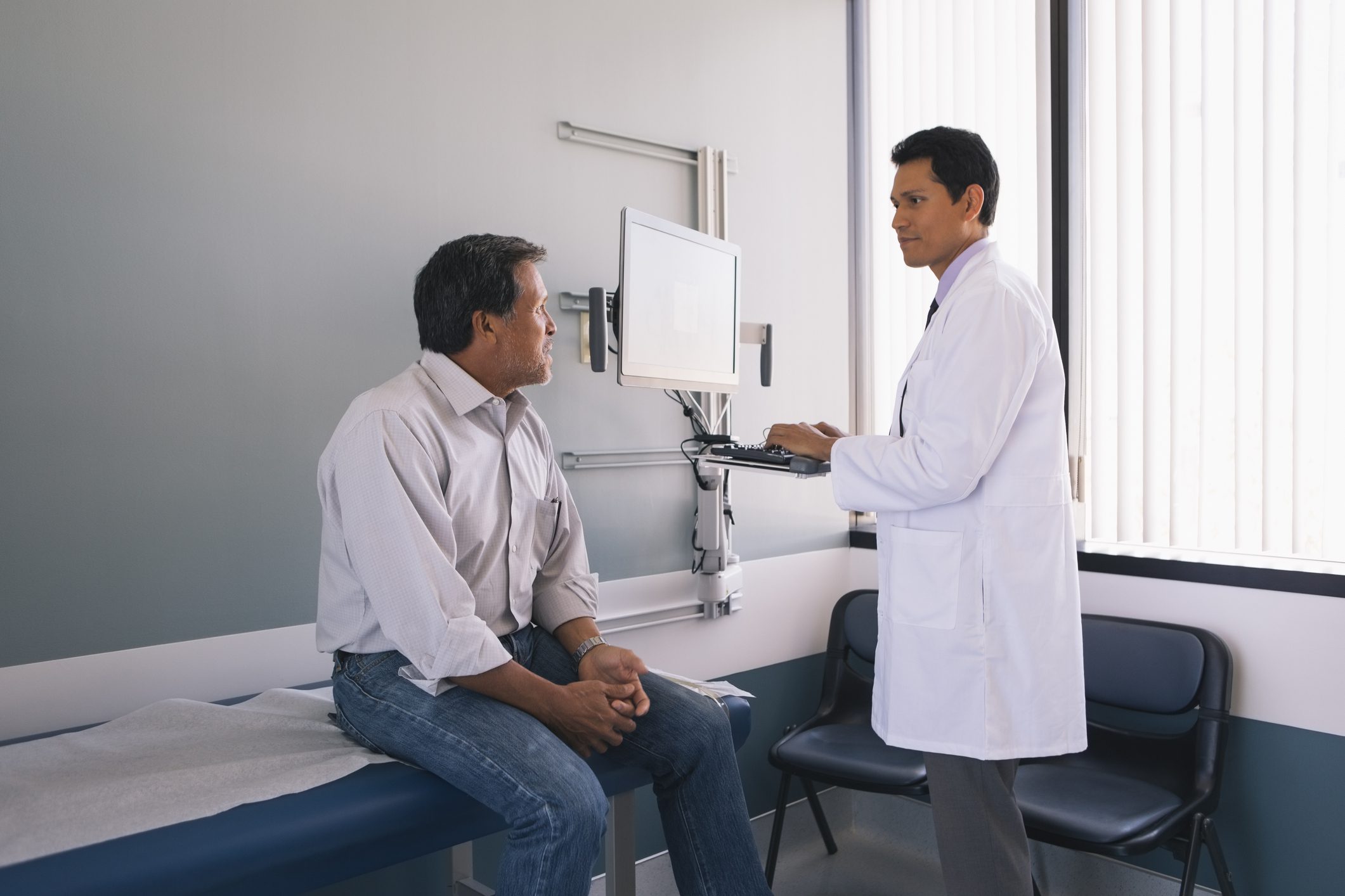 Male doctor working on computer while discussing with senior male patient in hospital